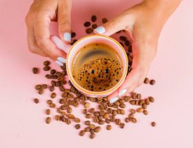 mains tenant une tasse de café entourée de grains de café sur fond rose