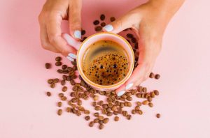 mains tenant une tasse de café entourée de grains de café sur fond rose