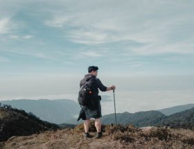 Solo hiker with backpack and walking stick looking at breathtaking mountain landscape under blue sky.