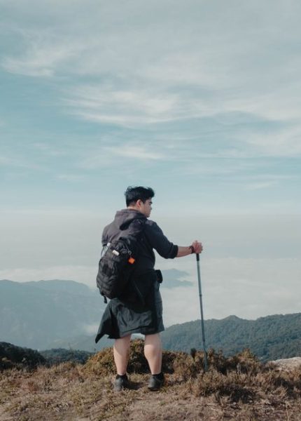 Solo hiker with backpack and walking stick looking at breathtaking mountain landscape under blue sky.
