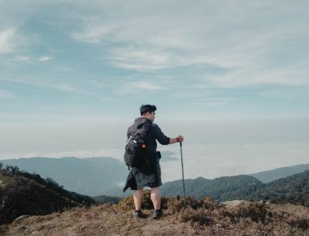 Solo hiker with backpack and walking stick looking at breathtaking mountain landscape under blue sky.