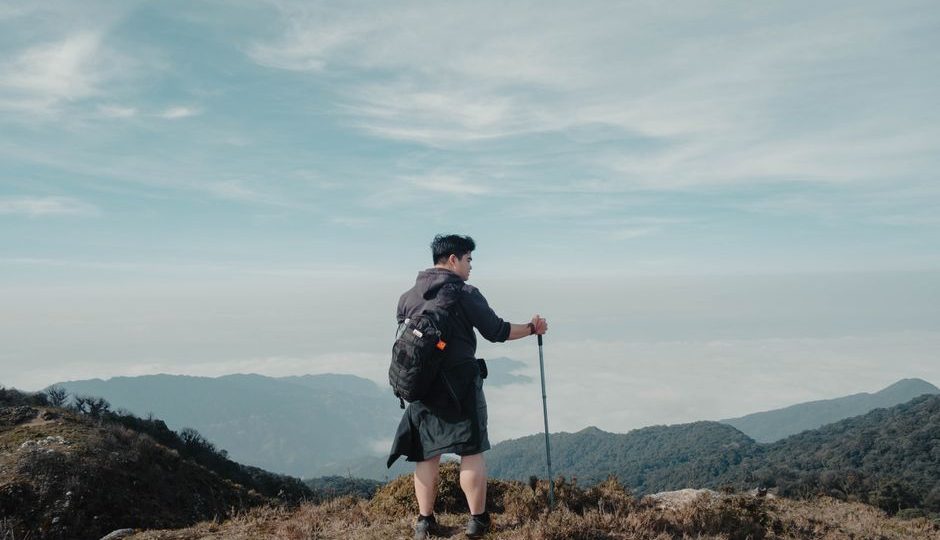 Solo hiker with backpack and walking stick looking at breathtaking mountain landscape under blue sky.